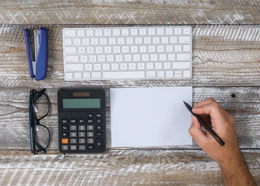 Hand notiert etwas neben Tastatur, Taschenrechner und Brille von oben
